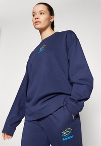 Felpa blu navy con polsi e orlo a coste, con un logo "Lotto" multicolore sul petto. Tessuto morbido e strutturato, vestibilità oversize.