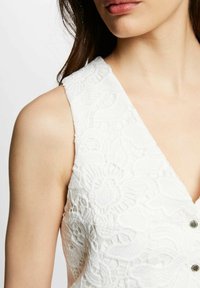 Top en dentelle blanche avec un décolleté en V profond, présentant des motifs floraux, un tissu texturé et des boutons décoratifs au centre. Détails de couture clairs visibles.