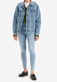 Chaqueta de mezclilla azul claro con dos bolsillos en el pecho, combinada con jeans ajustados azul medio y mocasines negros. Suéter verde texturizado por debajo.