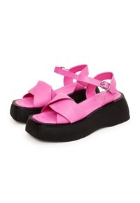 Sandales plateformes roses avec des brides croisées, fabriquées en matériau synthétique lisse, dotées d'une semelle texturée noire et de brides de cheville réglables.
