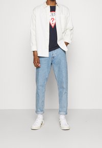 Camicia bianca a maniche lunghe, t-shirt grafica blu navy con logo a forma di cuore, jeans azzurri chiari e sneakers bianche. Cuffs arrotolati sui jeans; stile casual.