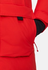 Giacca isolante rossa con superficie testurizzata, dettagli neri, tasca laterale e cordoncino regolabile per una vestibilità sicura.