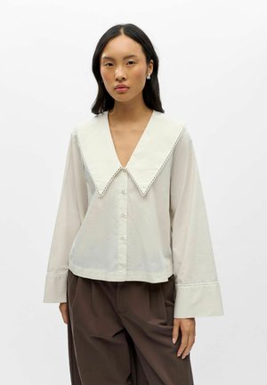 Femme portant un chemisier crème avec un grand col pointu et de larges poignets, associé à un pantalon marron ample sur un fond blanc uni.