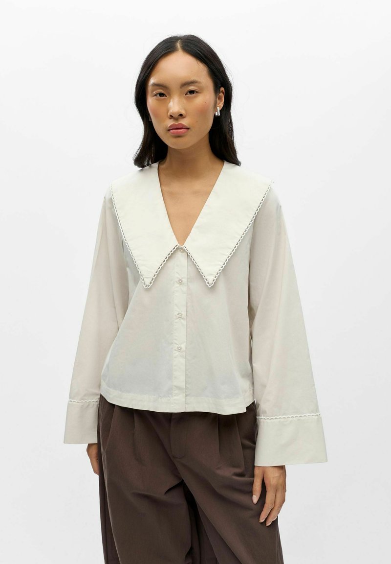 Femme portant un chemisier crème avec un grand col pointu et de larges poignets, associé à un pantalon marron ample sur un fond blanc uni.