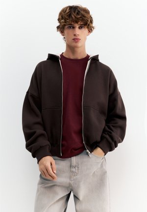 Mlada oseba z kodrastimi lasmi, oblečena v temno rjavo jopa z zadrgo čez bordo majico in svetlo sive kavbojke, stoji pred enobarvnim ozadjem.