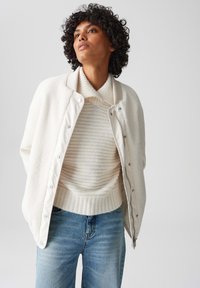 Witte fleecejas over een crème en witte gestreepte trui. Lichtblauwe denimjeans. Zachte texturen, relaxte pasvorm en drukknoopdetails.