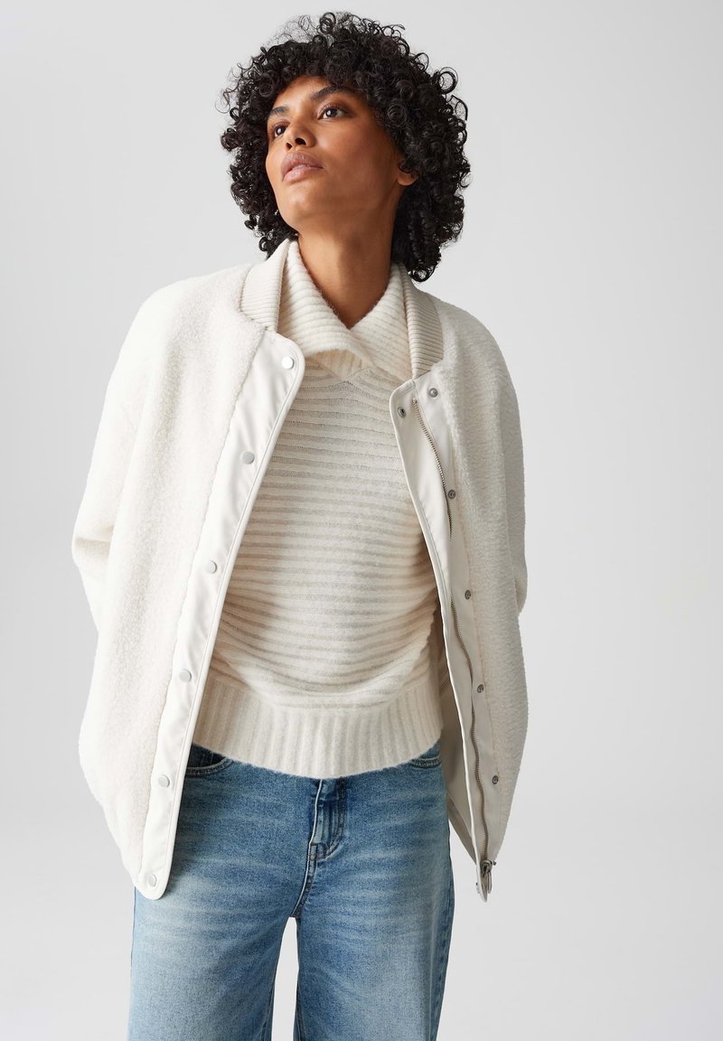 Witte fleecejas over een crème en witte gestreepte trui. Lichtblauwe denimjeans. Zachte texturen, relaxte pasvorm en drukknoopdetails.
