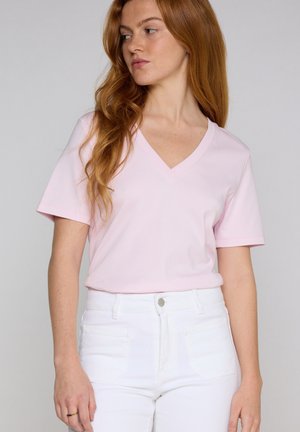 Femme aux longs cheveux roux porte un t-shirt rose clair à col en V et un jean blanc taille haute, regardant légèrement vers sa gauche sur un fond uni.