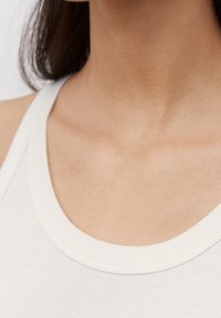 Gros plan sur le cou et le haut de la poitrine d'une personne portant un haut blanc sans manches avec des cheveux foncés tombant derrière l’épaule.