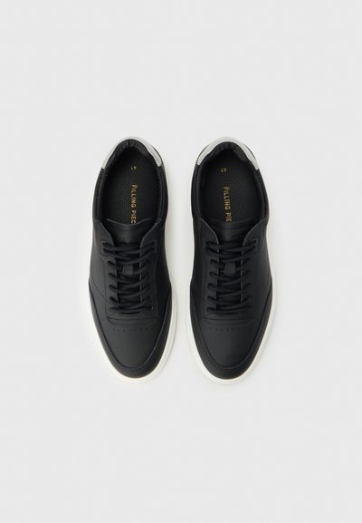 Ζευγάρι μαύρα sneakers χαμηλού ύψους με λευκές σόλες και μαύρα κορδόνια, εμφανιζόμενα από πάνω σε ένα απλό ανοιχτό γκρι φόντο.