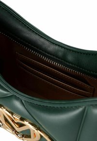 Interior de un bolso de mano verde de cuero que muestra un compartimento con cremallera marrón y ranuras para tarjetas, con detalles de hardware dorado en el borde exterior.