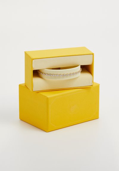 Une boîte à bijoux jaune contenant un bracelet en argent orné d'une rangée de strass transparents et scintillants sur un coussin beige.