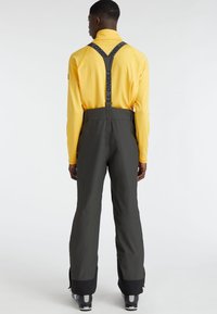 Pantalones de esquí resistentes al agua en gris oscuro con tirantes negros ajustables; llevados sobre una camiseta de manga larga brillante en color amarillo, con una textura suave.