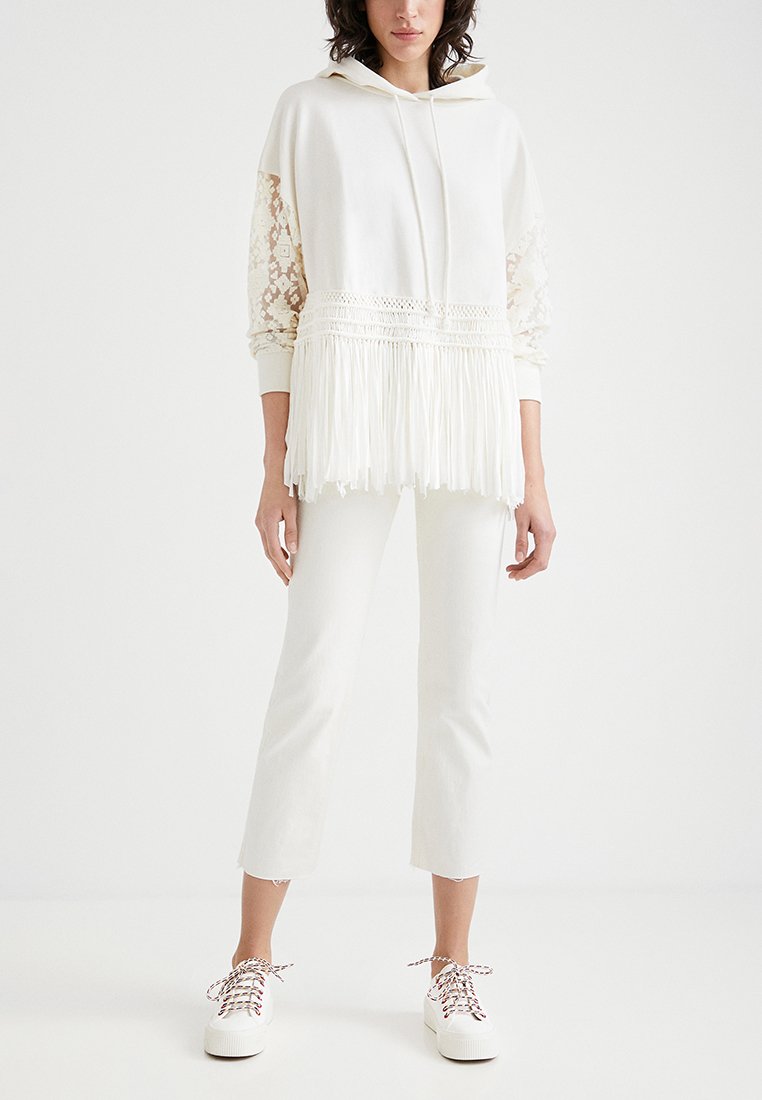 Felpa bianca con cappuccio e dettagli in pizzo, orlo a frange e maniche lunghe. Abbinata a jeans bianchi a vita alta e scarpe bianche con lacci colorati.