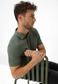 Grøn kortærmet polo-shirt i struktureret stof, slim fit, båret af en person siddende på en matchende grøn metalstol med vandrette lameller.
