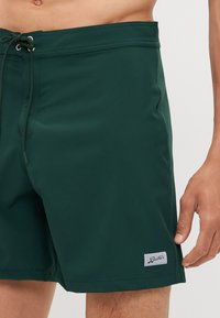Mørkegrønne badebukser laget av glatt stoff, med strikk i livet og en liten hvit logo-lapp på den nedre siden.