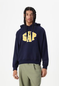 Hanorac albastru marin cu logo galben "GAP" pe față, dotat cu buzunar de tip kangaroo și glugă cu șnur. Material neted, croială lejeră.