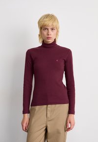 Maglione a collo alto color bordeaux con maniche lunghe e texture a costine. Presenta un piccolo logo sul petto. Abbastanza con pantaloni beige.