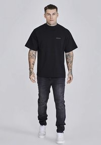 Juoda per didelio dydžio trumpomis rankovėmis marškinėliai su mažu logotipu. Derinami su tamsiomis džinsų kelnėmis ir baltais sportbačiais. Modelis stovi tiesiai.