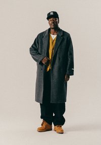 Homme noir portant une casquette noire, un manteau gris, un cardigan jaune, un pantalon noir et des bottes beiges, se tenant contre un fond clair et uni.