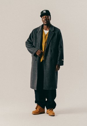 Schwarzer Mann trägt eine schwarze Mütze, einen grauen Überzieher, eine gelbe Strickjacke, eine schwarze Hose und braune Stiefel und steht vor einem einfarbigen hellen Hintergrund.