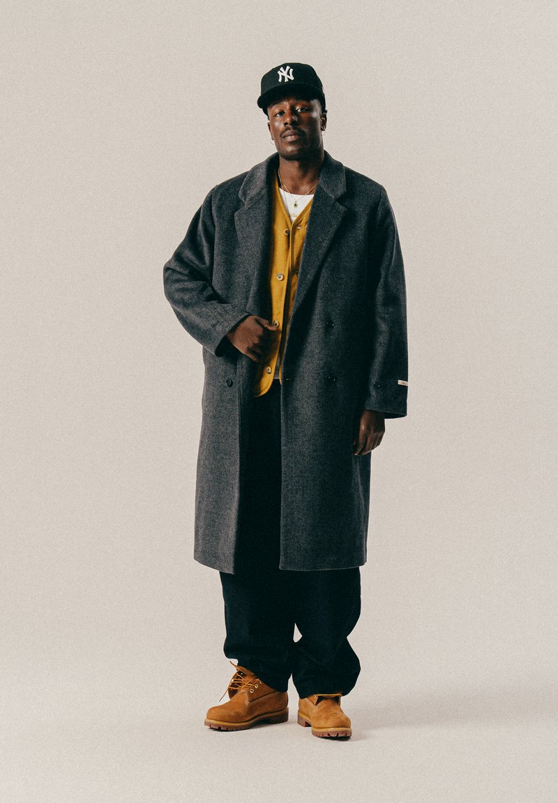 Homme noir portant une casquette noire, un manteau gris, un cardigan jaune, un pantalon noir et des bottes beiges, se tenant contre un fond clair et uni.