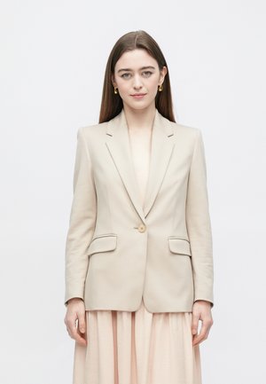 Beiger Blazer mit einem Knopf, Reverskragen und zwei Vorderentaschen. Hergestellt aus glattem Stoff, kombiniert mit einem hellrosa Kleid.