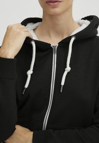 Crna zip-up dukserica napravljena od mekog materijala, s bijelim slojem od flisa, vrpcom s čvorastim krajevima i srebrnim patentnim zatvaračem.
