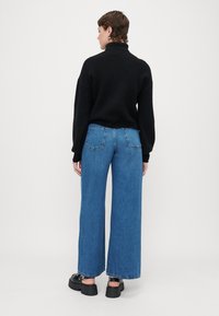 Suéter preto de malha com gola alta, combinado com jeans de algodão azul de perna larga, com bolsos traseiros e bainhas desfiadas. Sapatos pretos de plataforma.