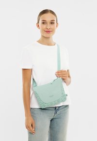 Kvinde iført hvid t-shirt og jeans holder en lys grøn crossbody taske med geometrisk mønster og lynlås, stående mod hvid baggrund.