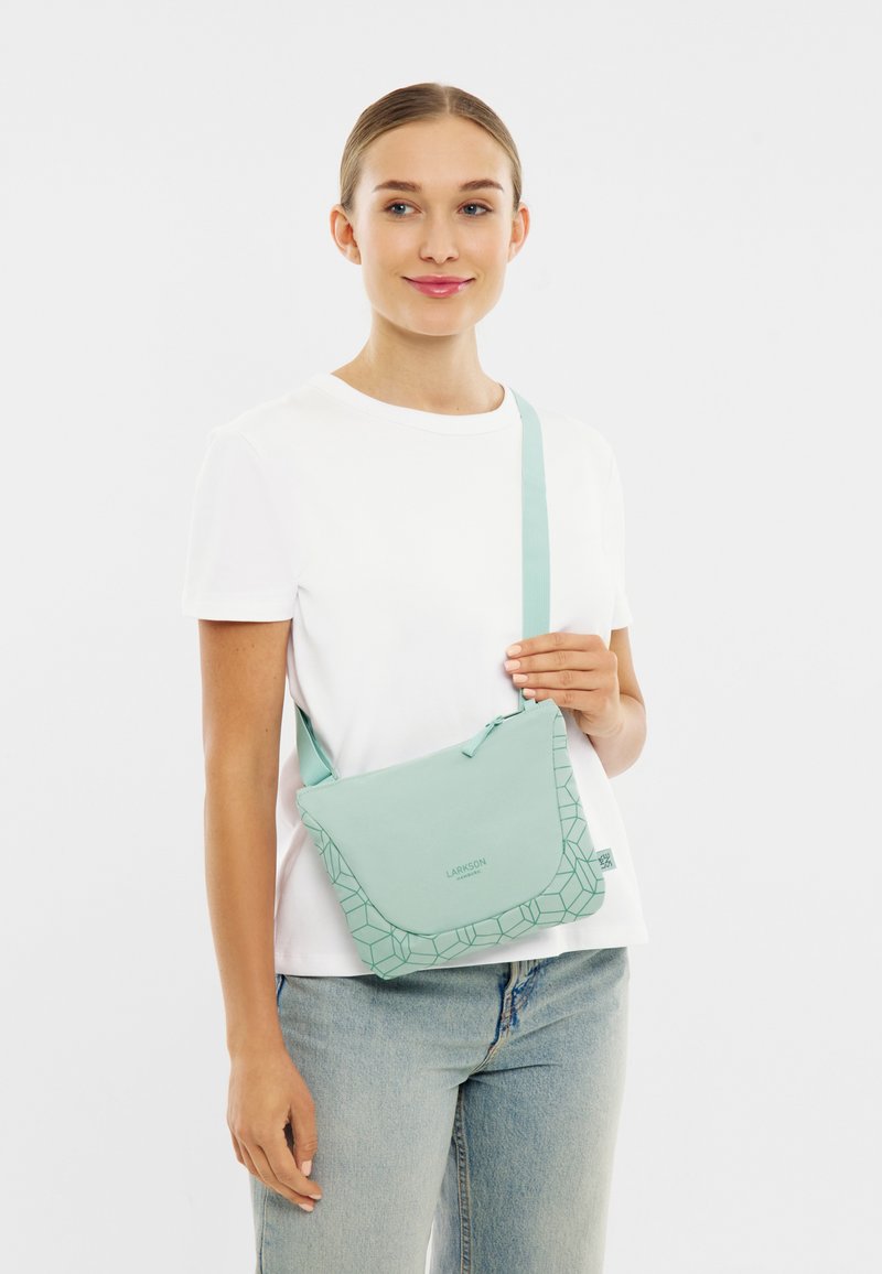Kvinde iført hvid t-shirt og jeans holder en lys grøn crossbody taske med geometrisk mønster og lynlås, stående mod hvid baggrund.