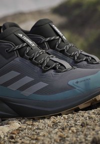Zapatillas de senderismo de color gris oscuro con material Gore-Tex, parte superior texturizada, puntera reforzada y acentos azules. Presenta cordones grises y el logo de tres rayas.