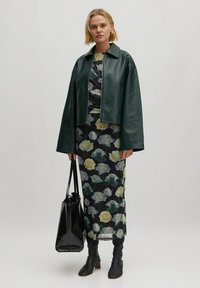 Zwart bloemenjurk met pale gele rozen, gecombineerd met een kort groen leren jasje. Model houdt een glanzende zwarte tote bag vast en draagt zwarte enkellaarsjes.