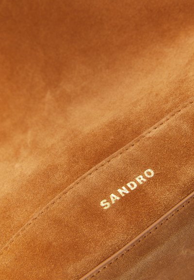 Lähikuva ruskeasta suede-kankaasta, jossa on ommeltu sauma ja kultainen "SANDRO"-logo painettuna pinnalle.