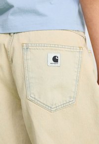 Ljusfärgade denimbyxor med en rektangulär ficka som har en svart och vit logotypfläck. Tyget har en subtil textur.