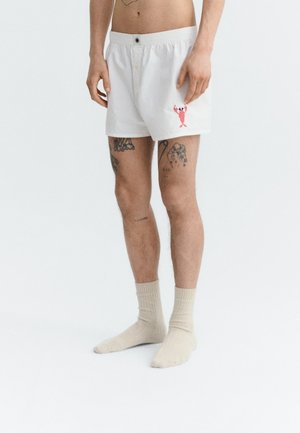 Mężczyzna w białych bokserkach z małym czerwonym motywem homara na lewej nogawce, beżowych skarpetkach i widocznymi tatuażami na obu nogach.