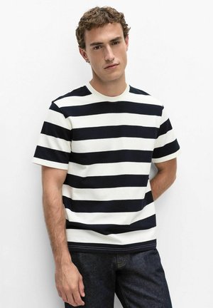 Jeune homme aux cheveux courts et bouclés portant une chemise à manches courtes rayée noire et blanche et un jean foncé, debout sur un fond blanc uni.