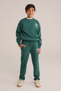 Groene sweatshirt en bijpassende joggers van zacht materiaal. De sweatshirt heeft bedrukte witte tekst; de joggers hebben zijzakken en elastische boorden.