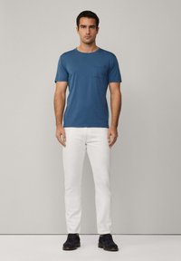 Modré bavlněné tričko s kapsou na prsou, kombinované s bílými slim-fit džíny. Model má tmavé boty a stojí na neutrálním pozadí.