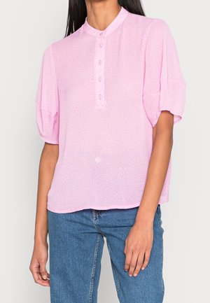 Femme portant un chemisier rose clair à manches courtes avec des boutons et un jean bleu, debout devant un fond clair uni.