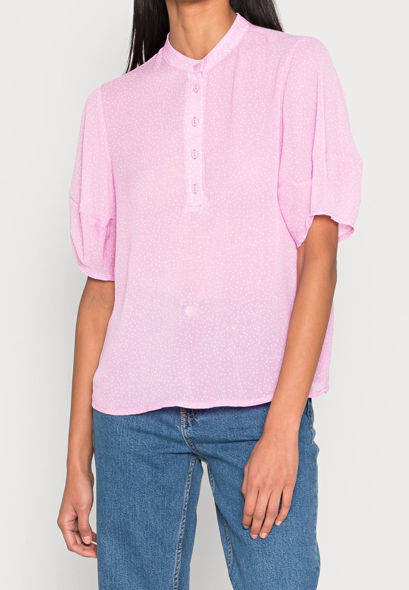 Femme portant un chemisier rose clair à manches courtes avec des boutons et un jean bleu, debout devant un fond clair uni.