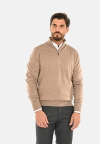 Maglione beige con zip al collo realizzato in tessuto morbido, caratterizzato da una trama sottile, polsini a costine e una vestibilità aderente, abbinato a pantaloni grigio scuro.