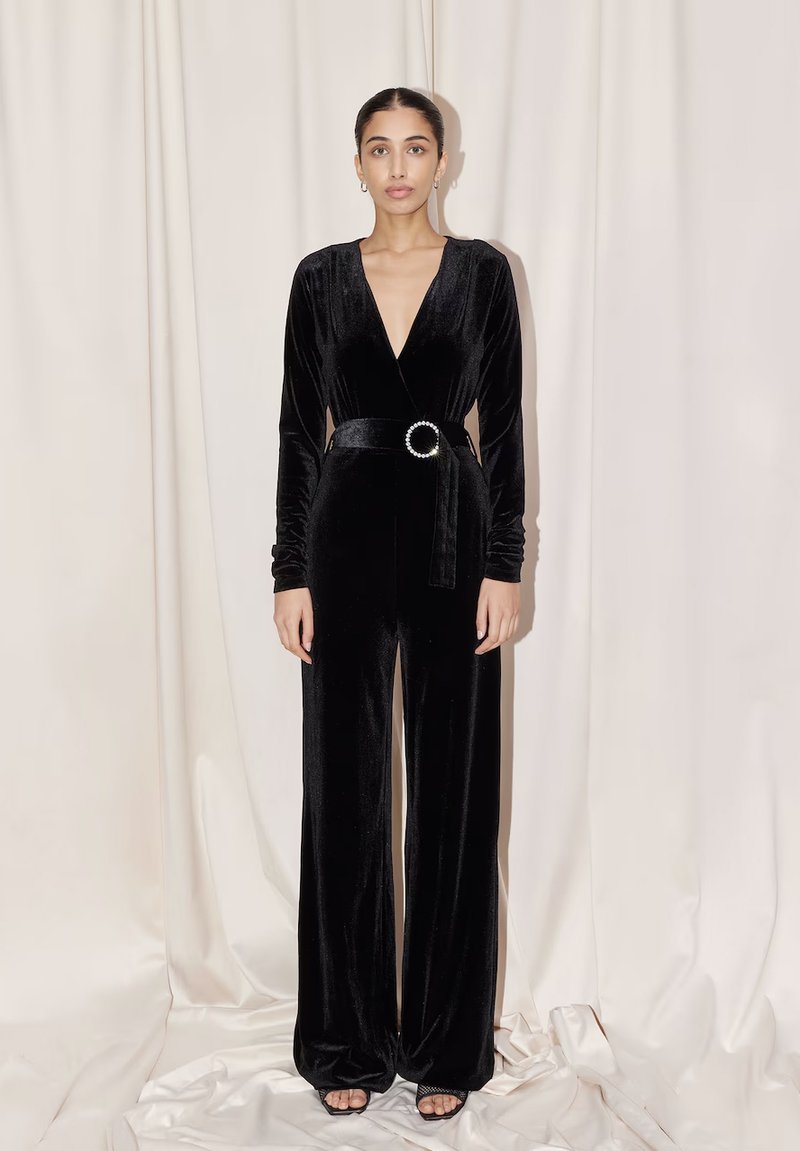 Jumpsuit in velluto nero con maniche lunghe, scollatura a V profonda, vita con cintura con un accento circolare e pantaloni a gamba larga. Tessuto morbido.