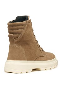 Botte chelsea en suède marron avec une surface texturée, bout rond et semelle en caoutchouc blanc présentant un motif à crampons. Détails de laçage et col rembourré.