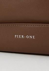 Bolso de cuero sintético marrón con una textura suave, que presenta el logo en relieve "PIER ONE" en blanco. Detalle de costuras visibles.