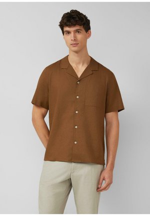 Junger Mann mit kurzen lockigen Haaren, der ein braunes kurzärmeliges Hemd und hellbeige Hose trägt, vor einem schlichten grauen Hintergrund steht.