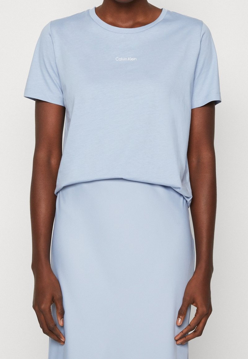 Nainen, joka pukeutunut vaaleansiniseen Calvin Kleinin lyhythihaiseen t-paitaan, joka on tungettu sopivaan vaaleansiniseen hameeseen, kuvattuna hartioista lantioihin.