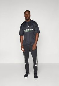 Juoda sportinė marškinėliai su žalia „ADIDAS“ logotipu ir atitinkamos juodos treniruočių kelnės su trim baltomis šoninėmis juostelėmis, dėvimi su juodais sportbačiais.