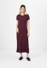 Bordowa sukienka maxi z krótkimi rękawami, okrągłym dekoltem, bocznymi rozcięciami i gładką fakturą. Noszona w połączeniu z czarnymi butami, na białym tle.