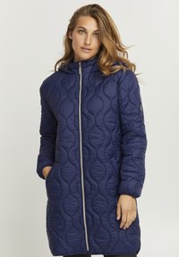 Cappotto lungo imbottito blu navy con chiusura a zip, cappuccio e tasche laterali. Presenta un motivo a onda testurizzato e polsini elasticizzati.